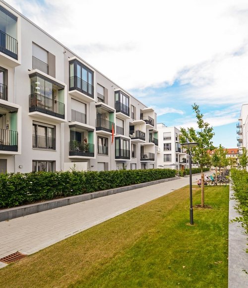 Eine moderne Wohnanlage mit mehreren weißen Gebäuden, die jeweils Balkone haben. Zwischen den Gebäuden verläuft ein gepflasterter Weg, der von gepflegten grünen Hecken und Rasenflächen gesäumt wird. Auf dem Weg sind einige Fahrräder abgestellt, und es stehen Laternen in der Landschaft. Der Himmel ist teilweise bewölkt und es ist Tageslicht zu sehen.