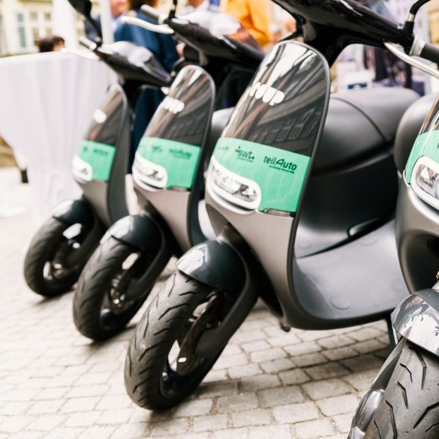 Vier schwarze E-Scooter stehen in einer Reihe, die vorderen Teile mit einer grünen Lackierung und den Aufschriften "powered by swt" und "teilAuto" versehen. Die Scooterräder sind sichtbar, und im Hintergrund sind verschwommene Menschen und eine gedeckte Fläche zu sehen. Die aufgeräumte Umgebung ist mit Steinpflaster gestaltet.