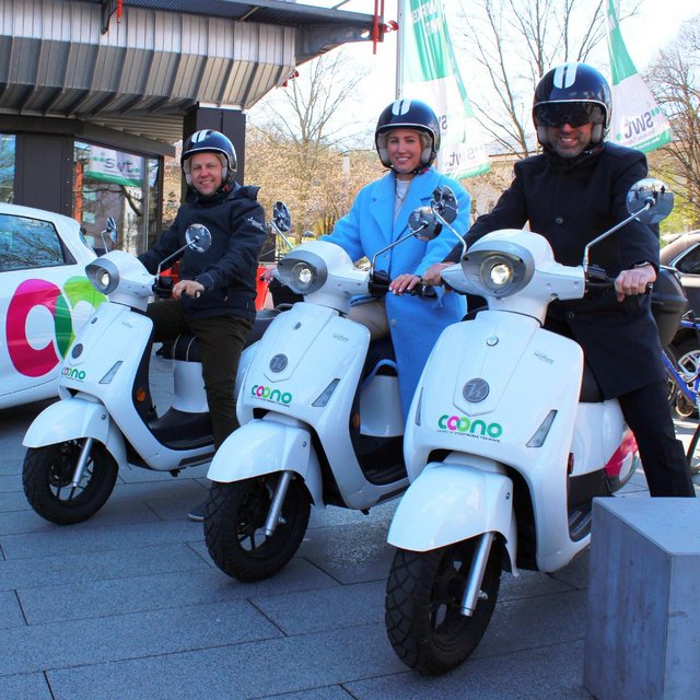 Drei Personen stehen auf weißen motorisierten Scootern, die mit einem Logo und Schriftzug versehen sind. Zwei Personen tragen Helme, während die dritte Person einen blauen Mantel und eine Sonnenbrille trägt. Im Hintergrund ist ein Fahrzeug mit einem Logo zu sehen, das ebenfalls zur gleichen Marke gehört, und einige Bäume sind sichtbar. Es ist ein sonniger Tag, und die Szenerie wirkt freundlich und lebhaft.
