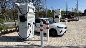 Eine weiße Elektroautoladestation mit zwei Ladepunkten steht in einem modernen urbanen Umfeld. Ein weißer PKW, der an einer der Ladestationen angeschlossen ist, ist im Vordergrund zu sehen. Im Hintergrund befinden sich andere Ladestationen, Straßenlaternen und eine Bushaltestelle. Die Umgebung ist grün mit Bäumen und Pflanzbeeten, und es gibt asphaltierte Wege und Pflastersteine auf dem Boden.