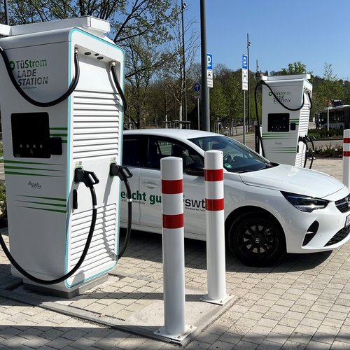Eine weiße Elektroautoladestation mit zwei Ladepunkten steht in einem modernen urbanen Umfeld. Ein weißer PKW, der an einer der Ladestationen angeschlossen ist, ist im Vordergrund zu sehen. Im Hintergrund befinden sich andere Ladestationen, Straßenlaternen und eine Bushaltestelle. Die Umgebung ist grün mit Bäumen und Pflanzbeeten, und es gibt asphaltierte Wege und Pflastersteine auf dem Boden.