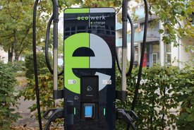 Eine Elektro-Ladestation steht an einer Straße, umgeben von Bäumen und Sträuchern. Die Station hat eine große, abgerundete Vorderseite mit einem modernen Design, das teilweise in Schwarz und Grün gehalten ist. Auf der Vorderseite sind das Wort "eco werk" und "e-charge" prominent abgebildet. Unterhalb des Displays befinden sich verschiedene Schaltflächen und zwei Steckdosen mit Handgriffen. Im Hintergrund sind Wohngebäude zu sehen, mit Fenstern und Balkonen.