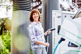 Eine Frau steht an einer Ladestation für Elektrofahrzeuge und hält ein Ladekabel in der Hand. Sie lächelt und trägt ein kariertes Hemd. Im Hintergrund sind Bäume und eine andere Säule der Ladestation sichtbar.