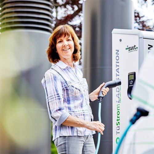 Eine Frau steht an einer Ladestation für Elektrofahrzeuge und hält ein Ladekabel in der Hand. Sie lächelt und trägt ein kariertes Hemd. Im Hintergrund sind Bäume und eine andere Säule der Ladestation sichtbar.