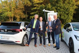 Eine Gruppe von vier Männern steht an einer Ladestation für Elektroautos. Sie halten ein Ladekabel in den Händen, während ein schwarzes und ein weißes Elektrofahrzeug in der Nähe geparkt sind. Im Hintergrund sind Bäume zu sehen, die die Szene umgeben. Die Männer tragen formelle Kleidung und lächeln in die Kamera.