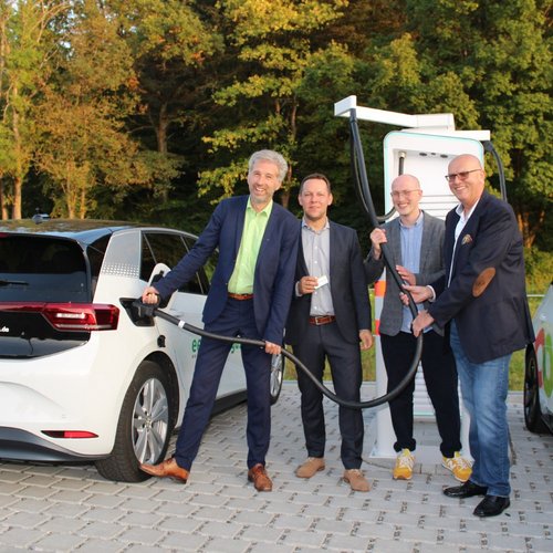 Eine Gruppe von vier Männern steht an einer Ladestation für Elektroautos. Sie halten ein Ladekabel in den Händen, während ein schwarzes und ein weißes Elektrofahrzeug in der Nähe geparkt sind. Im Hintergrund sind Bäume zu sehen, die die Szene umgeben. Die Männer tragen formelle Kleidung und lächeln in die Kamera.