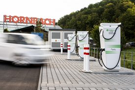 Eine unscharfe weiße Fahrzeugbewegung auf einem Parkplatz mit mehreren Elektroauto-Ladestationen. Im Hintergrund ist ein großes Schild mit der Aufschrift "HORNBACH" zu sehen. Die Ladestationen sind weiß mit grünen und schwarzen Aufschriften und haben Kabel, die zum Laden von Elektroautos verwendet werden können. Um die Ladestationen stehen rote und weiße Poller, und im Hintergrund sind Bäume und ein weiteres Gebäude sichtbar.