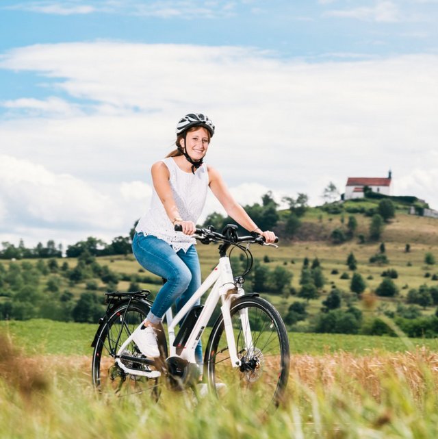 Eine Frau fährt auf einem weißen Fahrrad durch ein Feld. Sie trägt einen Helm und ein weißes Oberteil sowie Jeans. Im Hintergrund ist ein sanfter Hügel mit einer kleinen, weißen Gebäude zu sehen, und der Himmel ist sonnig mit einigen Wolken.