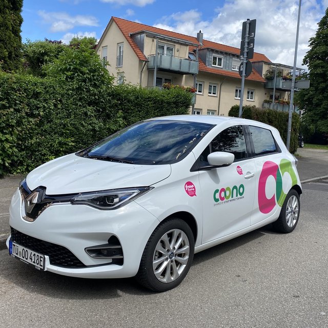 Ein weißer Renault ZOE ist auf einer Straße geparkt. Das Auto hat ein auffälliges Design mit bunten Logos und Texten auf der Seite, die "coono" und "free" zeigen. Im Hintergrund sind grüne Sträucher und einige Wohngebäude zu sehen. Der Himmel ist blau mit ein paar Wolken.