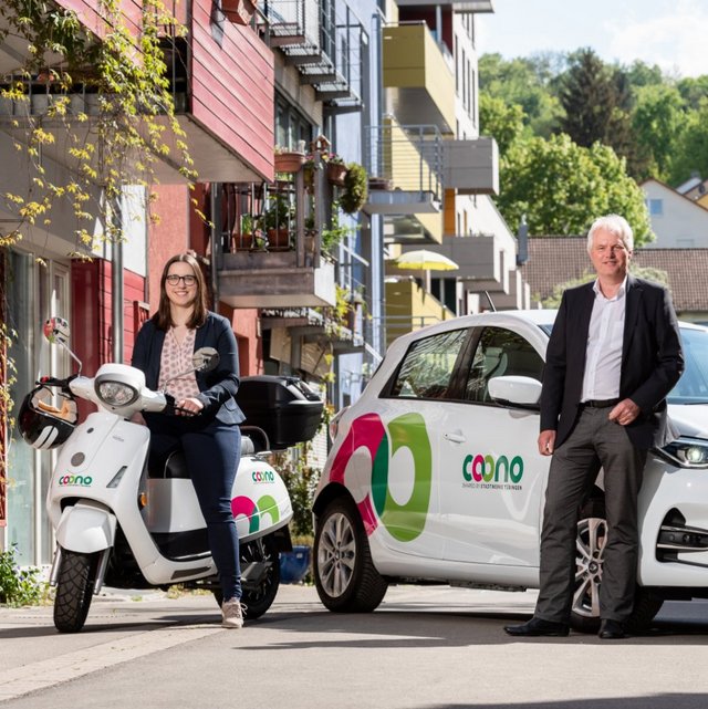 Eine Frau sitzt auf einem weißen Elektroroller und trägt eine schicke Bluse sowie eine dunkle Blazerjacke. Neben ihr steht ein Mann in einem Anzug, der lächelt. Im Hintergrund sind mehrstöckige Wohngebäude mit Balkonen und grünen Pflanzen zu sehen. Ein weißes Elektroauto, das ein Logo mit bunten Kreisen trägt, parkt neben dem Roller. Es ist ein sonniger Tag in einer urbanen Umgebung.