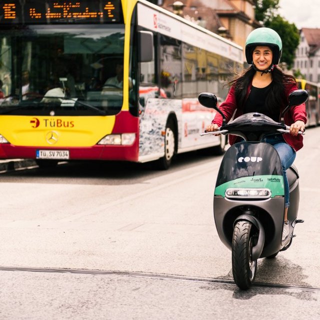 Eine Frau fährt auf einem Elektroroller in einer Stadtstraße. Sie trägt einen grünen Helm und ein rotes Oberteil. Im Hintergrund sind ein Linienbus und mehrere Autos zu sehen, während auf beiden Seiten des Straßenbildes Gebäude mit unterschiedlichen Farben und Stilen stehen. Die Frau lächelt und scheint Spaß beim Fahren zu haben.