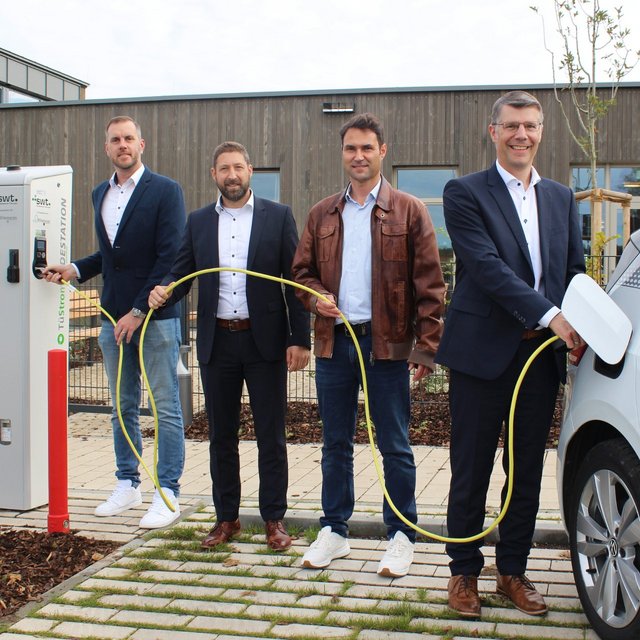 Vier Männer stehen an einer Ladestation für Elektrofahrzeuge. Der erste Mann links hält einen Stecker, der zweite Mann hat leichten Abstand und schaut zur Kamera, der dritte Mann trägt eine braune Jacke und steht in der Mitte, während der vierte Mann rechts einen Stecker in ein Auto steckt. Das Elektrofahrzeug ist an der Ladestation angeschlossen, und im Hintergrund ist ein modernes Gebäude zu sehen. Die Umgebung ist mit Pflanzen und einem gepflasterten Weg gestaltet.