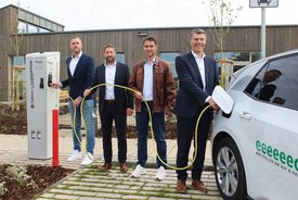 Vier Männer stehen an einer Ladestation für Elektrofahrzeuge. Der erste Mann links hält einen Stecker, der zweite Mann hat leichten Abstand und schaut zur Kamera, der dritte Mann trägt eine braune Jacke und steht in der Mitte, während der vierte Mann rechts einen Stecker in ein Auto steckt. Das Elektrofahrzeug ist an der Ladestation angeschlossen, und im Hintergrund ist ein modernes Gebäude zu sehen. Die Umgebung ist mit Pflanzen und einem gepflasterten Weg gestaltet.