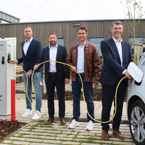 Vier Männer stehen an einer Ladestation für Elektrofahrzeuge. Der erste Mann links hält einen Stecker, der zweite Mann hat leichten Abstand und schaut zur Kamera, der dritte Mann trägt eine braune Jacke und steht in der Mitte, während der vierte Mann rechts einen Stecker in ein Auto steckt. Das Elektrofahrzeug ist an der Ladestation angeschlossen, und im Hintergrund ist ein modernes Gebäude zu sehen. Die Umgebung ist mit Pflanzen und einem gepflasterten Weg gestaltet.