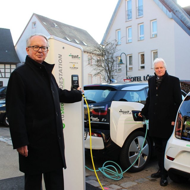 Zwei Männer in schwarzen Mänteln stehen neben Elektroautos, die an einer Ladestation angeschlossen sind. Der Mann auf der linken Seite hält den Stecker der Ladestation in der Hand, während er in die Kamera lächelt. Das Auto auf der linken Seite ist ein BMW i3, das auf der rechten Seite ein Volkswagen e-up!. Im Hintergrund sind einige Gebäude und Bäume sichtbar.