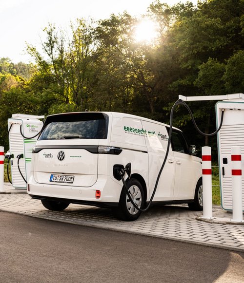 Eine weiße Elektrofahrzeug-Ladeeinrichter steht an einer modernen Ladestation in einer landschaftlich reizvollen Umgebung mit Bäumen im Hintergrund. Das Fahrzeug ist ein Volkswagen Bus, der mit einem Ladekabel an einer der zwei Ladeeinheiten angeschlossen ist. Neben der Ladestation sind rote und weiße Absperrpfosten zu sehen, und es gibt ein Gebäude im Hintergrund, das wahrscheinlich ebenfalls mit elektrischer Energie oder Infrastruktur verbunden ist. Der Himmel ist teilweise bewölkt, und die Sonne scheint durch die Bäume.