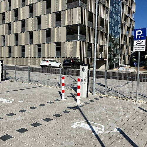 Ein Parkplatz mit gestreifter Pflasterung, auf dem weiße Markierungen für Parkplätze sichtbar sind. Im Hintergrund befindet sich ein modernes Gebäude mit einer Fassade aus senkrechten, perforierten Paneelen. Es gibt mehrere Parkschilder, darunter eines, das auf Ladevorgänge hinweist. Zwei Poller mit roten und weißen Streifen sperren den Zugang, und im Hintergrund sind mehrere Autos zu sehen.