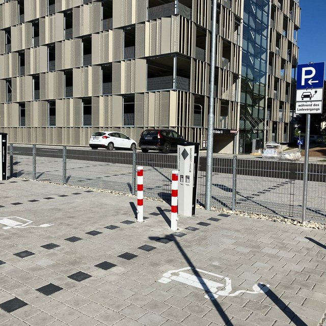 Ein Parkplatz mit gestreifter Pflasterung, auf dem weiße Markierungen für Parkplätze sichtbar sind. Im Hintergrund befindet sich ein modernes Gebäude mit einer Fassade aus senkrechten, perforierten Paneelen. Es gibt mehrere Parkschilder, darunter eines, das auf Ladevorgänge hinweist. Zwei Poller mit roten und weißen Streifen sperren den Zugang, und im Hintergrund sind mehrere Autos zu sehen.