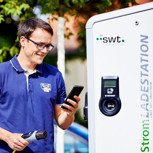 Ein junger Mann steht neben einer Ladestation für Elektroautos. Er trägt ein blaues Polo-Shirt und hält ein Smartphone in der rechten Hand. In der linken Hand hält er das Kabel der Ladestation. Der Hintergrund zeigt Bäume und andere Fahrzeuge. Der Mann lächelt und scheint auf das Smartphone zu schauen. Die Ladestation ist mit dem Aufdruck „Strom LADESTATION“ versehen.