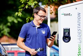 Ein junger Mann steht neben einer Ladestation für Elektroautos. Er trägt ein blaues Polo-Shirt und hält ein Smartphone in der rechten Hand. In der linken Hand hält er das Kabel der Ladestation. Der Hintergrund zeigt Bäume und andere Fahrzeuge. Der Mann lächelt und scheint auf das Smartphone zu schauen. Die Ladestation ist mit dem Aufdruck „Strom LADESTATION“ versehen.