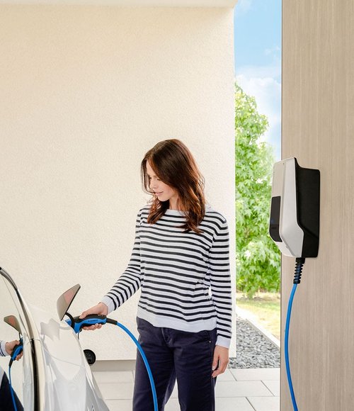 Eine Frau mit langen, braunen Haaren steht neben einem weißen Elektroauto und steckt das Ladekabel in den Ladeanschluss des Fahrzeugs. Sie trägt ein gestreiftes Oberteil in Schwarz und Weiß sowie dunkelblaue Hosen. Im Hintergrund ist eine Wand sowie eine grüne Pflanze sichtbar, und der Himmel ist blau mit einigen Wolken. An der Wand hängt eine Ladestation für das Elektroauto.