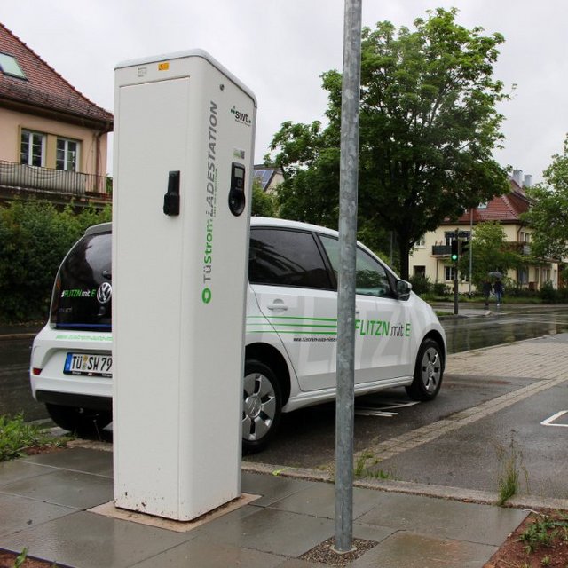 Eine elektrische Ladestation steht an einer Straße, neben der ein weißer VW-Dienstwagen mit der Aufschrift "FLITZN mit E" parkt. In der Nähe sind grüne Ampeln zu sehen, und die Straße ist nass von recentem Regen. Im Hintergrund sind einige Wohnhäuser und Bäume sichtbar, die der Szene eine grüne Umgebung verleihen. Auf dem Boden sind Markierungen für das Parken von Elektrofahrzeugen zu erkennen.