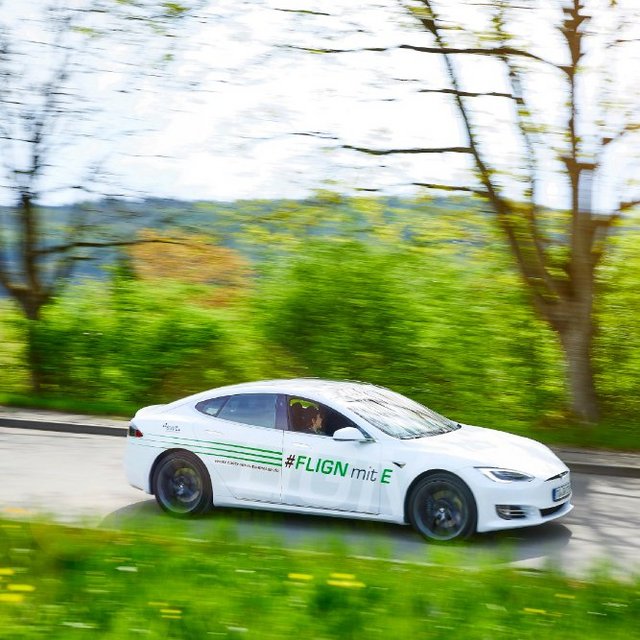 Ein weißes Elektroauto fährt schnell auf einer kurvigen Landstraße, umgeben von üppigem grünem Gras und Bäumen. Auf der Seite des Fahrzeugs steht der Hashtag "#FLIGNmitE". Der Autofahrer sitzt am Steuer und trägt eine Sonnenbrille. Das Bild vermittelt Bewegung und Dynamik durch die verschwommenen Hintergründe. Es ist ein sonniger Tag mit strahlendem Licht.