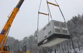 Ein Kran hebt einen großen, rechteckigen silbergrauen Container, der möglicherweise ein Generator oder ein klimatisiertes Gerät ist. Der Container schwebt in der Luft, während im Hintergrund schneebedeckte Bäume und eine winterliche Landschaft zu sehen sind. Der Kran hat eine leuchtend orange Farbe mit dem Schriftzug "Wiesbauer".