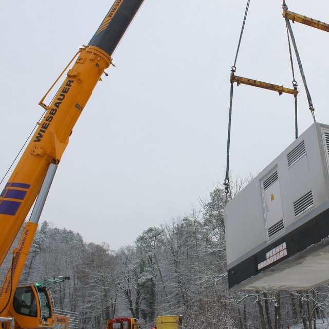 Ein Kran hebt einen großen, rechteckigen silbergrauen Container, der möglicherweise ein Generator oder ein klimatisiertes Gerät ist. Der Container schwebt in der Luft, während im Hintergrund schneebedeckte Bäume und eine winterliche Landschaft zu sehen sind. Der Kran hat eine leuchtend orange Farbe mit dem Schriftzug "Wiesbauer".
