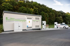 Eine moderne Ladestation für Elektrofahrzeuge steht an einer Straße. Die Station ist mit einem großen Logo versehen, das "TüStrom LADESTATION" zeigt, und hat den Hinweis "Schnelles Laden MIT 300 KW" auf der Wand. Neben der Station befinden sich zwei Ladestellen mit Kabeln, die in ein Elektrofahrzeug eingesteckt sind. Im Hintergrund sind grüne Bäume und ein klarer Himmel zu sehen. Ein weißes Elektrofahrzeug ist an der Station geparkt.