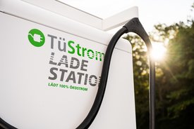 Eine Elektroauto-Ladestation mit dem Schriftzug "TüStrom LADE STATION". Der Text beschreibt, dass 100% Ökostrom geladen wird. Ein Kabel hängt an der Ladestation, und im Hintergrund ist Sonnenlicht, das durch Bäume scheint.