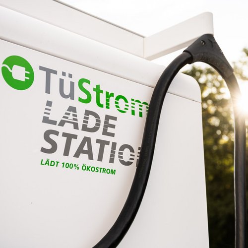 Eine Elektroauto-Ladestation mit dem Schriftzug "TüStrom LADE STATION". Der Text beschreibt, dass 100% Ökostrom geladen wird. Ein Kabel hängt an der Ladestation, und im Hintergrund ist Sonnenlicht, das durch Bäume scheint.