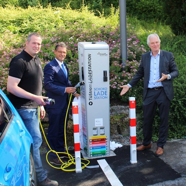 Drei Männer stehen an einer Ladestation für Elektrofahrzeuge, die an einer vielbefahrenen Straße steht. Die Ladestation ist mit bunten Kabeln und Schildern versehen, die auf die Nutzung hinweisen. Im Hintergrund sind Blumen und Bäume zu sehen, die die Umgebung grüner wirken lassen. Zwei der Männer halten Ladekabel in der Hand, während einer von ihnen mit der anderen Hand auf die Ladestation zeigt. Ein blauer und ein weißer Elektrofahrzeug stehen neben der Ladestation.