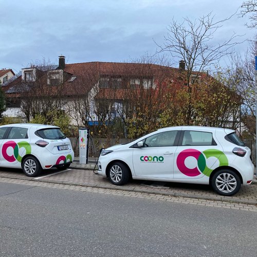Zwei weiße Elektroautos mit bunten Logos parken an einer Straße. Sie sind an einer Ladestation angeschlossen, die sich in der Nähe befindet. Im Hintergrund sind einige Häuser mit roten Dächern und Bäume zu sehen. Ein Parkschild steht ebenfalls an der Straße. Der Himmel ist bewölkt, und die Umgebung wirkt herbstlich.
