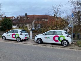 Zwei weiße Elektroautos mit bunten Logos parken an einer Straße. Sie sind an einer Ladestation angeschlossen, die sich in der Nähe befindet. Im Hintergrund sind einige Häuser mit roten Dächern und Bäume zu sehen. Ein Parkschild steht ebenfalls an der Straße. Der Himmel ist bewölkt, und die Umgebung wirkt herbstlich.