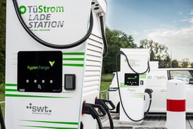 Zwei Ladestationen für Elektrofahrzeuge stehen vor einem weißen Fahrzeug. Die Stationen sind in Weiß mit grünen Akzenten gestaltet und tragen die Aufschrift "TuStrom LADESTATION" sowie das Logo "hypercharger". An den Stationen hängen Ladekabel, die bereit sind, in Elektrofahrzeuge eingesteckt zu werden. Im Hintergrund sind Bäume und ein klarer Himmel zu sehen. An einer der Ladestationen ist ein kleineres Gerät sichtbar, und eine Säule mit rotem und weißem Streifen ist ebenfalls im Bild.