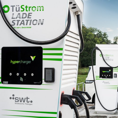 Zwei Ladestationen für Elektrofahrzeuge stehen vor einem weißen Fahrzeug. Die Stationen sind in Weiß mit grünen Akzenten gestaltet und tragen die Aufschrift "TuStrom LADESTATION" sowie das Logo "hypercharger". An den Stationen hängen Ladekabel, die bereit sind, in Elektrofahrzeuge eingesteckt zu werden. Im Hintergrund sind Bäume und ein klarer Himmel zu sehen. An einer der Ladestationen ist ein kleineres Gerät sichtbar, und eine Säule mit rotem und weißem Streifen ist ebenfalls im Bild.