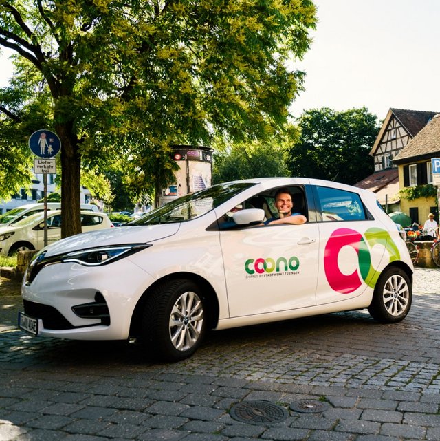 Ein weißes Elektroauto mit einem bunten Logo steht auf einer gepflasterten Straße. Der Fahrer, ein lächelnder Mann, ist am Steuer. Im Hintergrund sind mehrere Bäume und ein schönes, älteres Gebäude mit einem gedämpften gelben Anstrich zu sehen. Einige Personen sitzen auf einer Bank und unterhalten sich, während Fahrräder im Hintergrund abgestellt sind. Es ist ein sonniger Tag.