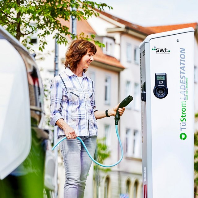 Eine Frau steht an einer Ladestation für Elektrofahrzeuge und hält ein Ladekabel in der Hand. Die Station ist hell und prägnant beschriftet mit „TüStrom LADES TATION“. Im Hintergrund sind einige Gebäude und Bäume zu sehen, die eine städtische Umgebung zeigen. Die Frau trägt ein kariertes Hemd und sieht freundlich aus, während sie sich auf das Laden des Fahrzeugs konzentriert. In der Nähe steht ein Elektroauto.