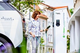 Eine Frau steht an einer Ladestation für Elektrofahrzeuge und hält ein Ladekabel in der Hand. Die Station ist hell und prägnant beschriftet mit „TüStrom LADES TATION“. Im Hintergrund sind einige Gebäude und Bäume zu sehen, die eine städtische Umgebung zeigen. Die Frau trägt ein kariertes Hemd und sieht freundlich aus, während sie sich auf das Laden des Fahrzeugs konzentriert. In der Nähe steht ein Elektroauto.