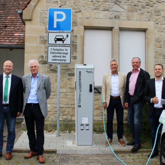 Eine Gruppe von fünf Männern steht vor einer Steinfassade mit einem geschlossenen Fenster. Neben ihnen befinden sich ein Ladestationen für Elektrofahrzeuge und Parkplatzschilder. Im Hintergrund ist ein Banner mit der Aufschrift „Wir wirren mit“ zu sehen. Ein Elektrofahrzeug ist am rechten Bildrand abgebildet.