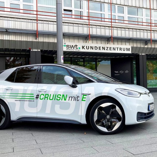 Ein weißes Elektroauto mit dem Schriftzug "#CRUISN mit E" steht vor einem modernen Gebäude mit großen Fenstern und einem Schild, das "swt. KUNDENZENTRUM" zeigt. Im Hintergrund sind grüne Bäume und zwei rote Pflanzgefäße sichtbar. Die Umgebung wirkt urban und einladend, mit einem gepflasterten Boden und einem Fahrradständer.